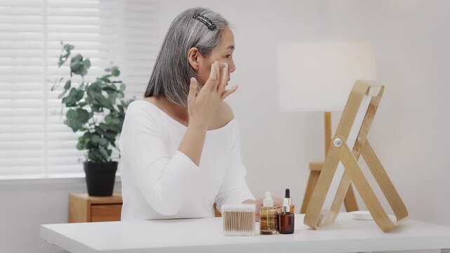 Asian mature woman with natural grey hair applies moisturizing facial cream to her cheek while looking in a mirror. Slow-motion shot.