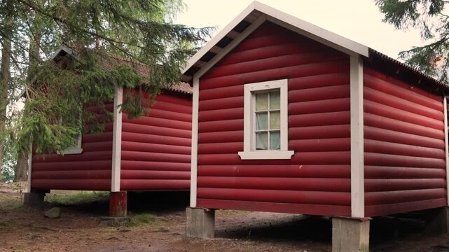 Traditional red wooden log cabin house stands elevated on concrete blocks among green pine trees, featuring white trim window and gabled roof in rustic Scandinavian forest setting.