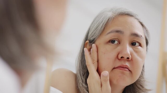 An Asian mature woman with grey hair looks in a mirror with concern while checking fine lines and aging skin.