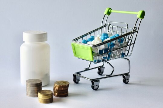 A white pill container, stacked money coins, and a miniature shopping cart filled with medical capsules against a white backdrop.