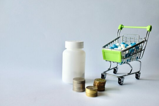 A white pill container, stacked money coins, and a miniature shopping cart filled with medical capsules against a white backdrop.