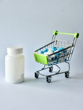 Miniature shopping cart filled with medical capsules and tablets placed next to a blank white medicine bottle, representing healthcare costs, pharmaceutical shopping, and medication expenses.