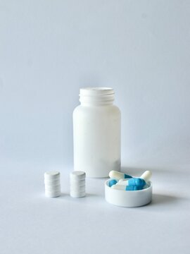 White pill bottle next to stacked round tablets and blue-white capsule medication in a plastic cap on a neutral background.