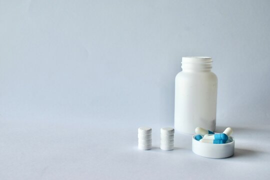 White pill bottle next to stacked round tablets and blue-white capsule medication in a plastic cap on a neutral background.