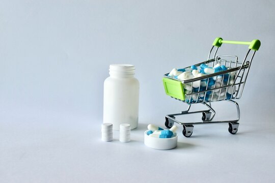 Miniature grocery cart overflowing with capsule medication set alongside a white pill bottle and neatly stacked tablets.