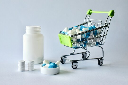 Miniature grocery cart overflowing with capsule medication set alongside a white pill bottle and neatly stacked tablets.