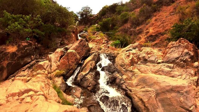 Slow motion view of a small waterfall trickling over golden and reddish rocks surrounded by natural vegetation near Usisya, Malawi.