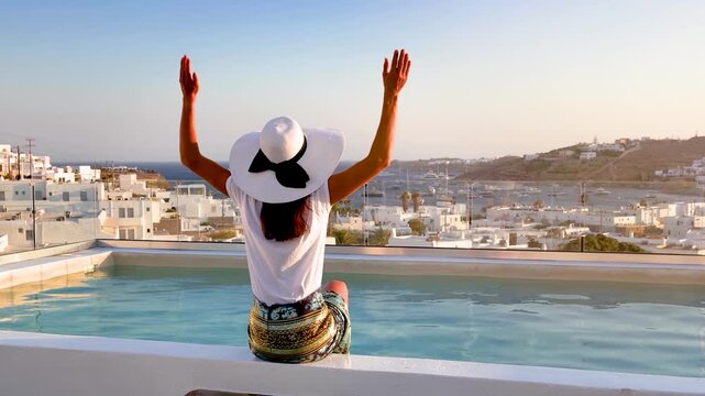 A happy woman sits by the swiming pool and looks at the island of Mykonos, Cyclades, Greece, during summer sunset time