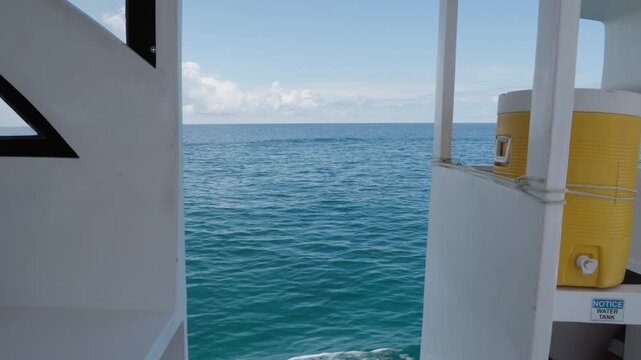 A steady side view from a moving boat frames turquoise Indian Ocean water, a foaming wake and open horizon between white deck panels and a yellow water dispenser in the Maldives.