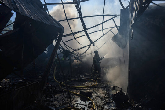 Firefighter in protective gear extinguishes a structural fire with a high-pressure fire hose amid dense smoke, flames, twisted metal debris, collapsed structures, and hazardous conditions.