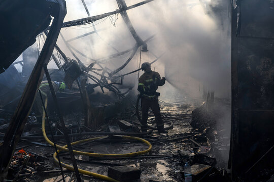 Firefighter in protective gear extinguishes a structural fire with a high-pressure fire hose amid dense smoke, flames, twisted metal debris, collapsed structures, and hazardous conditions.