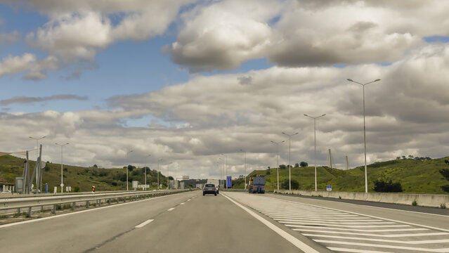 Vehicles travel along a wide motorway surrounded by rolling green hills under dramatic cloudy skies.