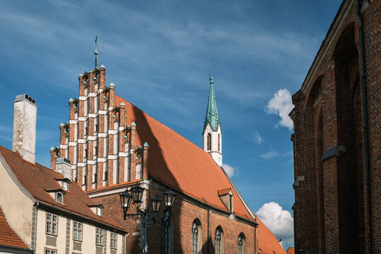 Wide low-angle view showing the Baltic Gothic stepped gable facade of St John Church framed by historic buildings in Riga, Latvia.