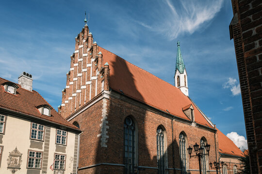 Wide low-angle view showing the Baltic Gothic stepped gable facade of St John Church framed by historic buildings in Riga, Latvia.