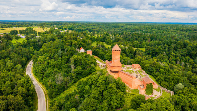 Aerial vista of medieval red brick Turaida Castle and tower surrounded by dense green forest and a winding rural road in Sigulda, Gauja National Park, Latvia.