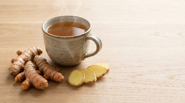 Steaming cup of herbal tea with fresh turmeric root and sliced turmeric on wooden surface