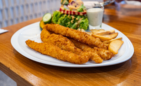 Plate of crispy fish fingers with potato wedges and a fresh salad served on a wooden table.