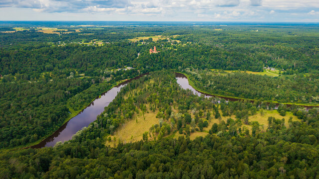 drone panorama showing the winding Gauja River and Turaida Castle emerging above green forest canopy under sunny sky in Latvia