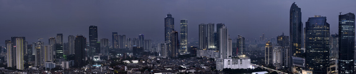 Obraz premium Panoramic wide-angle night view of a sprawling modern city skyline with illuminated skyscrapers under a dark, overcast twilight sky. Cityscape background