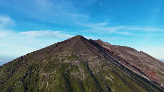 Aerial view of the volcanic peak of Mount Agung with rocky slopes and green vegetation under a clear blue sky in Rendang, Bali, Indonesia.