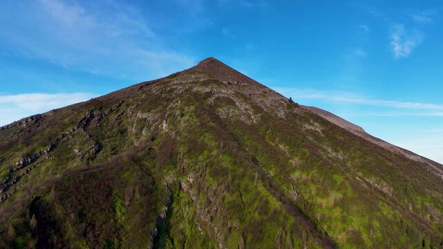 Aerial view of the volcanic slopes of Mount Agung with lush green vegetation and rocky terrain under a clear blue sky in Rendang, Bali, Indonesia.
