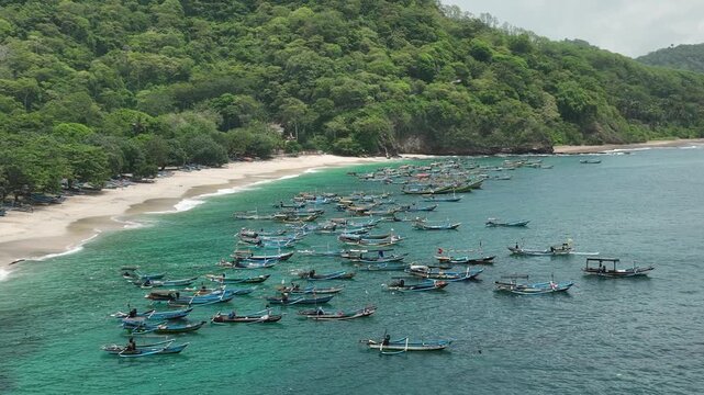 Aerial view of Papuma Beach with numerous traditional fishing boats anchored in the turquoise water along the sandy coastline in Jember, East Java, Indonesia.
