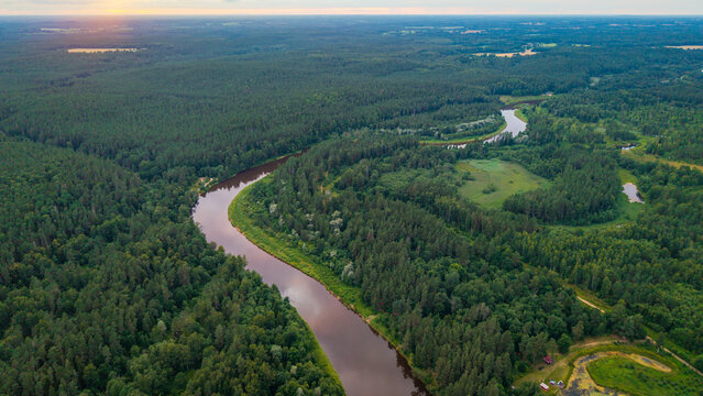 Aerial sunset view over Gauja River winding through dense green forest towards glowing horizon in Sigulda, Latvia