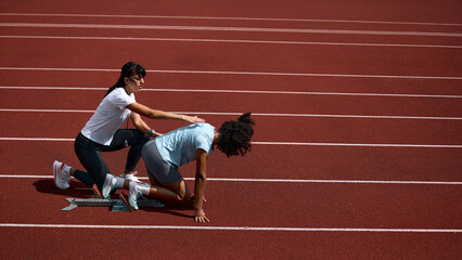 Naklejka premium Adult female coach correcting teenage runner crouched starting position on track. Concept of hands on training assistance, athlete safety instruction, summer sport mentorship.