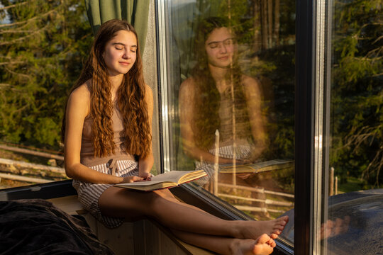 Teenage Girl Relaxing with Book by Sunlit Window. Teenage girl sits barefoot by a large window with an open book on her lap, resting with closed eyes in warm sunlight.