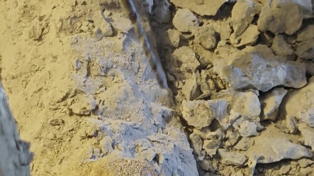 a construction worker holding a heavy-duty rotary hammer drill, breaking through concrete near a brick wall. Dust clouds and pulverized stone fragments spray upward during indoor renovation