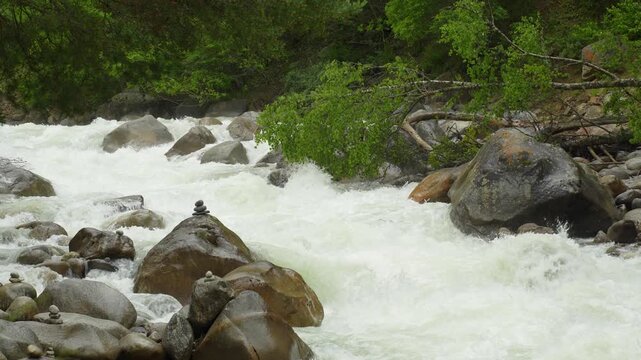 A dynamic section of a mountain river: foaming water rushes between massive, rounded boulders, curving around them and forming eddies. In the foreground are neatly stacked pyramids of stones.
