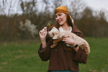 Fototapeta premium Joyful woman in a casual jacket outdoors holds a goat kid, offering a dandelion, smiling warmly in a green field, capturing a gentle countryside lifestyle and affectionate animal interaction