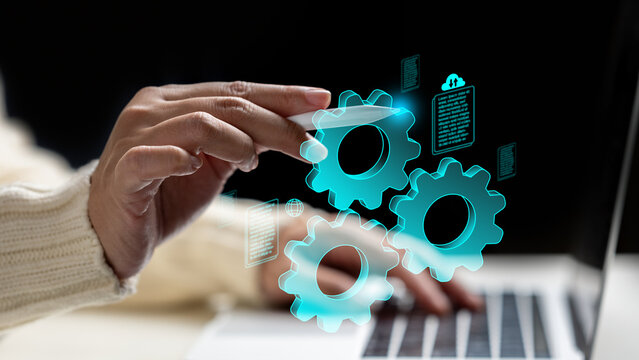 A woman hand using a stylus to interact with glowing blue virtual gears and document icons over a laptop, representing digital automation and workflow management. Data and automation concept