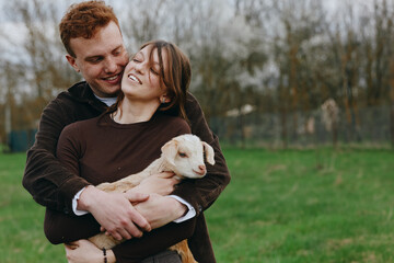 Naklejka premium Joyful couple outdoors with a gentle goat kid, embracing and smiling, in a park setting, showcasing affectionate lifestyle moment, warm connection, and playful pet interaction