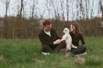 Naklejka premium Casual outdoor lifestyle with a cheerful couple sitting in a field, playing with a goat kid, warm afternoon light, relaxed smiles, gentle interaction, natural setting, candid moment of joy and