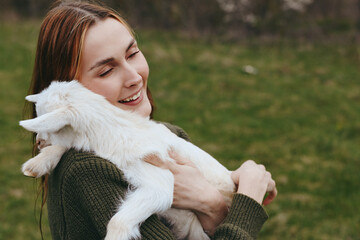Naklejka premium Lifestyle woman goat kid outdoors affection cuddle nature happiness photography. A candid scene of a smiling woman holding a goat kid in a grassy field, celebrating connection and gentle bonding.