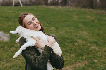 Naklejka premium Joyful woman outdoors hugs a fluffy goat kid, sharing a tender moment in a grassy field, casual sweater, peaceful countryside mood, candid lifestyle scene, gentle interaction with animals.
