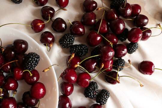 Fresh ripe sweet cherries and dark blackberries scattered on a white crumpled linen tablecloth with natural sunlight shadows.