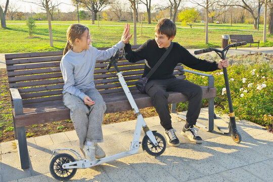 Brother and sister high fiving on a wooden bench with kick scooters in an autumn park. Happy smiling children friendship scene
