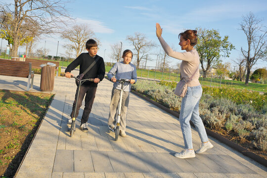 Mother high fiving her son and daughter on scooters in a park. Happy active family weekend