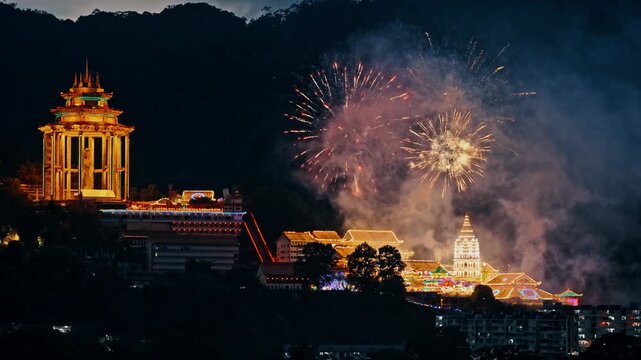 Wide static night shot of bright golden and red fireworks exploding over the brightly illuminated Kek Lok Si Temple complex and the towering Guan Yin statue in Penang.