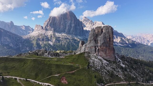 Aerial drone view of the Cinque Torri rock formations with the imposing Tofane massif in the background, surrounded by alpine meadows, rocky slopes, hiking trails, and scattered clouds