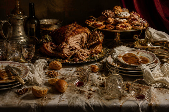 Wide still-life of overloaded banquet table after long meal, torn roast, stacked plates, scattered bread, tipped wine glasses and pastries, gluttony mood