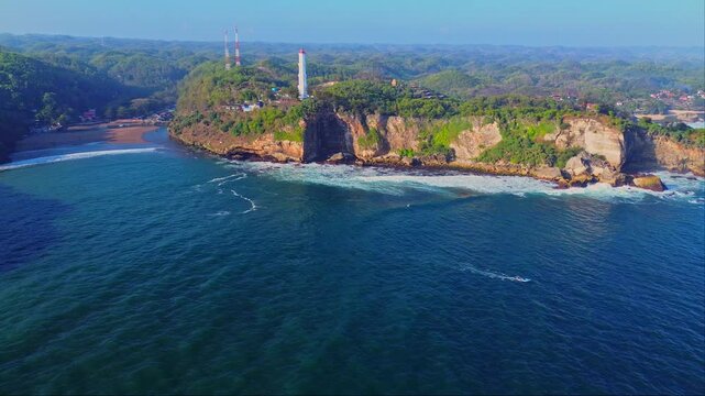 Aerial view of Baron Beach with its iconic lighthouse perched on rugged cliffs overlooking the blue ocean waves in Yogyakarta, Special Region of Yog, Indonesia.
