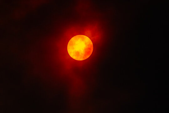 View of a glowing sun partially obscured by dark, hazy clouds, creating a dramatic, fiery red and orange appearance in the night sky in Colorado Springs, CO, United States.