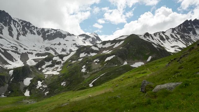 A panoramic view of a mountain range. In the foreground, a grassy slope with rocks and wildflowers; in the middle zone, steep ridges with patches of snow; in the background, massive peaks with exposed