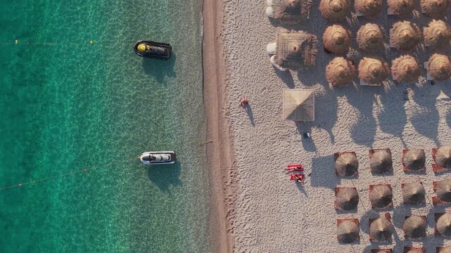 Aerial view of Llamani Beach with turquoise water, jet skis, and rows of straw umbrellas on the sand in Himare, Vlore County, Albania.