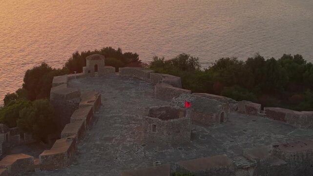 Aerial view of the historic Porto Palermo Castle with stone walls and an Albanian flag flying at sunset in Porto Palermo, Qarku i Vlores, Albania.