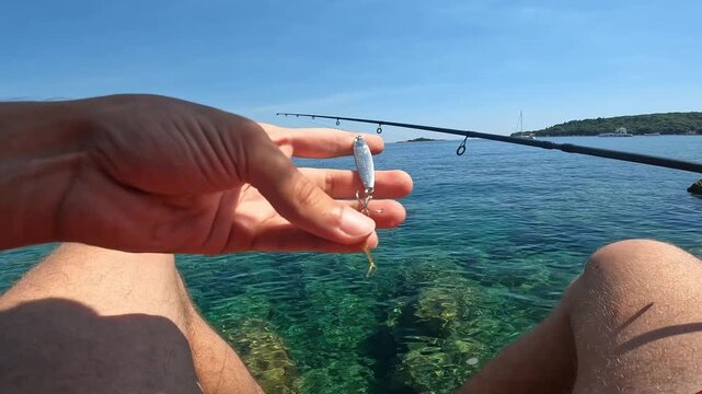 POV fisherman hand holding silver spoon lure with treble hook. Close up of fishing tackle gear against blue Mediterranean sea.