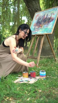 Beautiful woman artist in a dress painting on a canvas on an easel in nature by the river